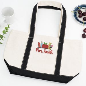 Canvas tote bag with black handles, embroidered apple and school supplies design, personalized with teacher name “Mrs. Smith,” styled with cup and cherries.
