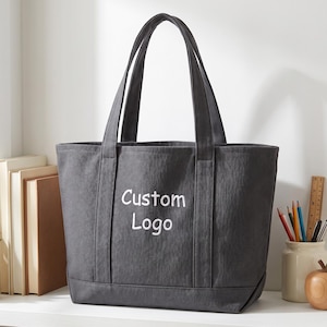 Custom logo tote bag in dark grey canvas with simple branding text, minimal workspace setup with books and stationery, ideal corporate merchandise