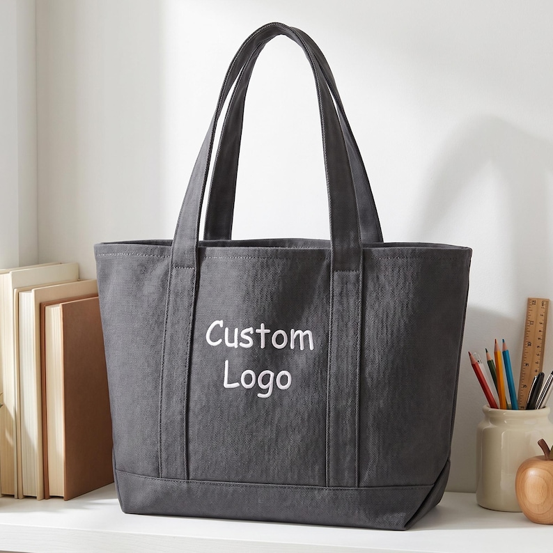 Custom logo tote bag in dark grey canvas with simple branding text, minimal workspace setup with books and stationery, ideal corporate merchandise