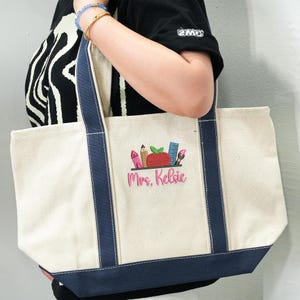 Large canvas tote bag with navy blue handles, embroidered school supplies and apple design, customized with teacher name “Mrs. Kelsie,” shown on model’s shoulder.