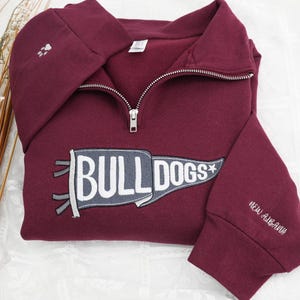 Puede incluir: Una sudadera burdeos con cremallera de un cuarto con un diseño de banderín "BULLDOGS" blanco y gris. El puño de la manga tiene bordado "NEW ALBANY" en blanco. Una huella de pata está bordada en la capucha.