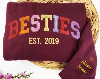 May include: Burgundy sweatshirt with the word "BESTIES" embroidered in rainbow colors and the text "EST. 2019" in gold. The sleeve has the names "Hunter" and "Scottie" embroidered in gold with a small heart.
