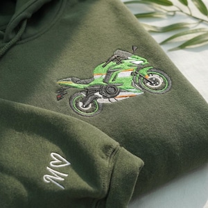 May include: An olive green hooded sweatshirt featuring an embroidered graphic of a green and white sport motorcycle on the chest. The sleeve displays white embroidered text that reads "MV" with a small heart symbol above the letters.