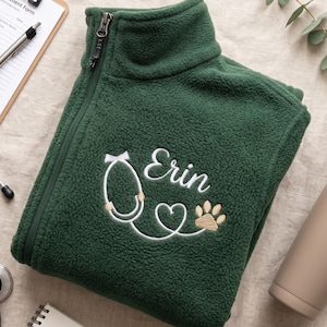 Peut inclure: Une veste polaire vert foncé à fermeture éclair latérale, ornée d'une broderie blanche représentant un stéthoscope, un cœur et une empreinte de patte. Le prénom Erin est brodé en lettres cursives blanches au-dessus du motif du stéthoscope.