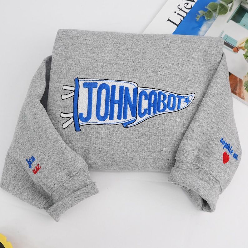 Puede incluir: Una sudadera gris con un bander&iacute;n azul y blanco que dice "JOHN CABOT". La manga tiene el texto "sophie m." y un coraz&oacute;n rojo. La otra manga tiene el texto "jca mac."