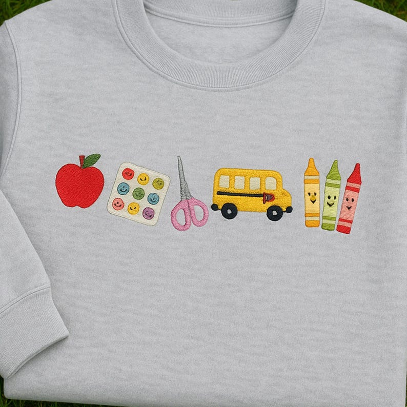 Folded grey sweatshirt with colorful embroidered school icons including apple, paint palette, scissors, school bus, and crayons – perfect back-to-school gift for teachers.