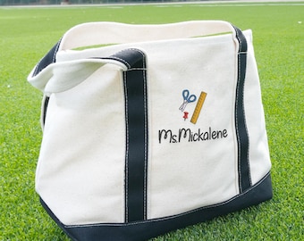 May include: Cream-colored tote bag with black trim and handles. The bag features an embroidered design of scissors, a ruler, and a push pin, with the name "Ms. Mickalene" below. The bag is sitting on green grass.