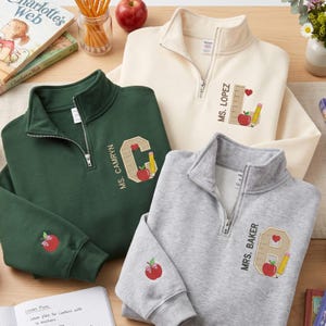 May include: Three quarter-zip sweatshirts in dark green, cream, and heather gray. Each features a personalized name, a large letter, a ruler, a pencil, and a red apple graphic. The sleeves are embroidered with a small red apple and a pink bow.
