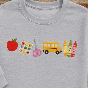 Light grey embroidered teacher sweatshirt featuring apple, smiley stickers, scissors, yellow bus, and crayons – fun and thoughtful gift for first day of school or Teacher Appreciation Week.