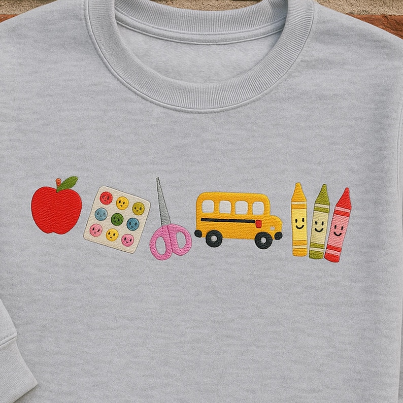 Light grey embroidered teacher sweatshirt featuring apple, smiley stickers, scissors, yellow bus, and crayons – fun and thoughtful gift for first day of school or Teacher Appreciation Week.