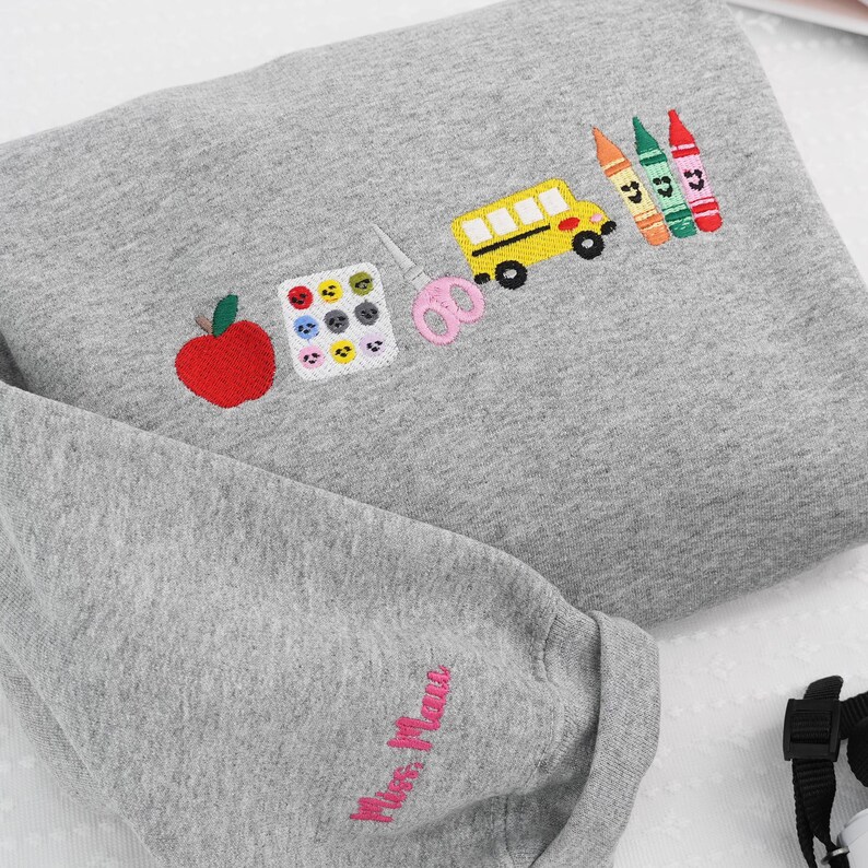 Personalized grey teacher sweatshirt with name embroidered on sleeve and school-themed design on chest – charming custom gift for new school year or classroom reveal.