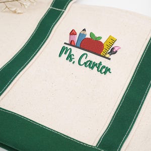 Teacher tote bag with green handles, embroidered school supplies and red apple design, customized with teacher name “Ms. Carter,” close-up view.