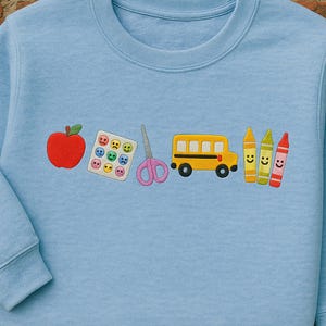 Light blue back-to-school sweatshirt with cute embroidered icons for teachers – apple, sticker chart, scissors, bus, and happy crayons – ideal gift for pre-k and elementary educators.