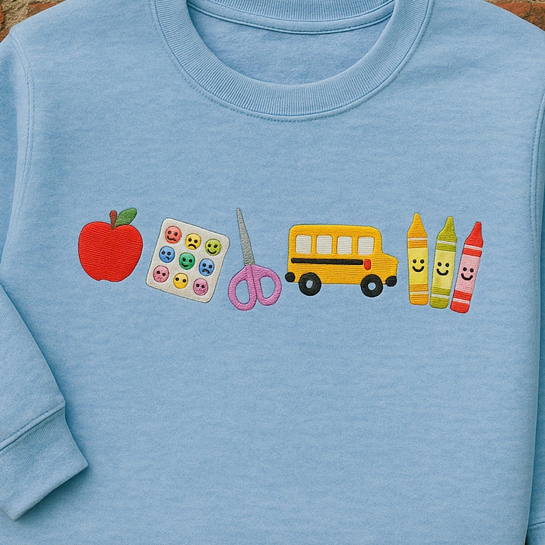 Light blue back-to-school sweatshirt with cute embroidered icons for teachers – apple, sticker chart, scissors, bus, and happy crayons – ideal gift for pre-k and elementary educators.