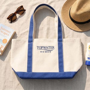 Custom logo tote bag for business branding with “Topwater Homes” print, beach lifestyle setting with summer accessories, promotional company tote
