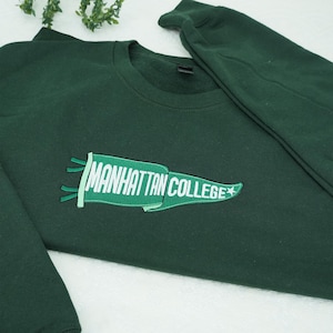 Puede incluir: Sudadera verde oscuro con la inscripci&oacute;n bordada "MANHATTAN COLLEGE" en una pancarta verde con texto blanco y una estrella blanca. La sudadera tiene cuello redondo y mangas largas.