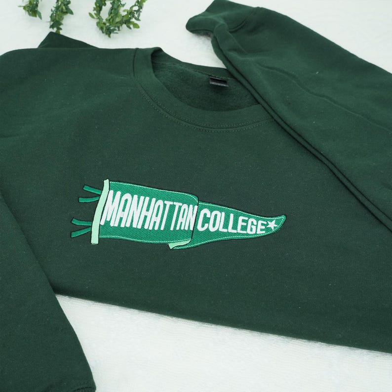 Puede incluir: Sudadera verde oscuro con la inscripci&oacute;n bordada "MANHATTAN COLLEGE" en una pancarta verde con texto blanco y una estrella blanca. La sudadera tiene cuello redondo y mangas largas.