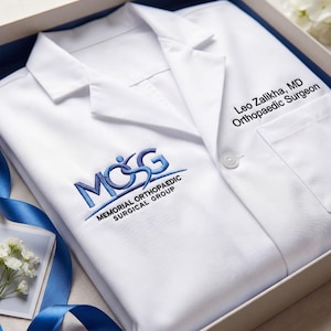 Puede incluir: Una bata de laboratorio médica blanca con el logotipo bordado en azul del Memorial Orthopaedic Surgical Group. El nombre "Leo Zalikha, MD Orthopaedic Surgeon" está impreso en la bata. La bata está cuidadosamente doblada en una caja de regalo con una cinta azul.