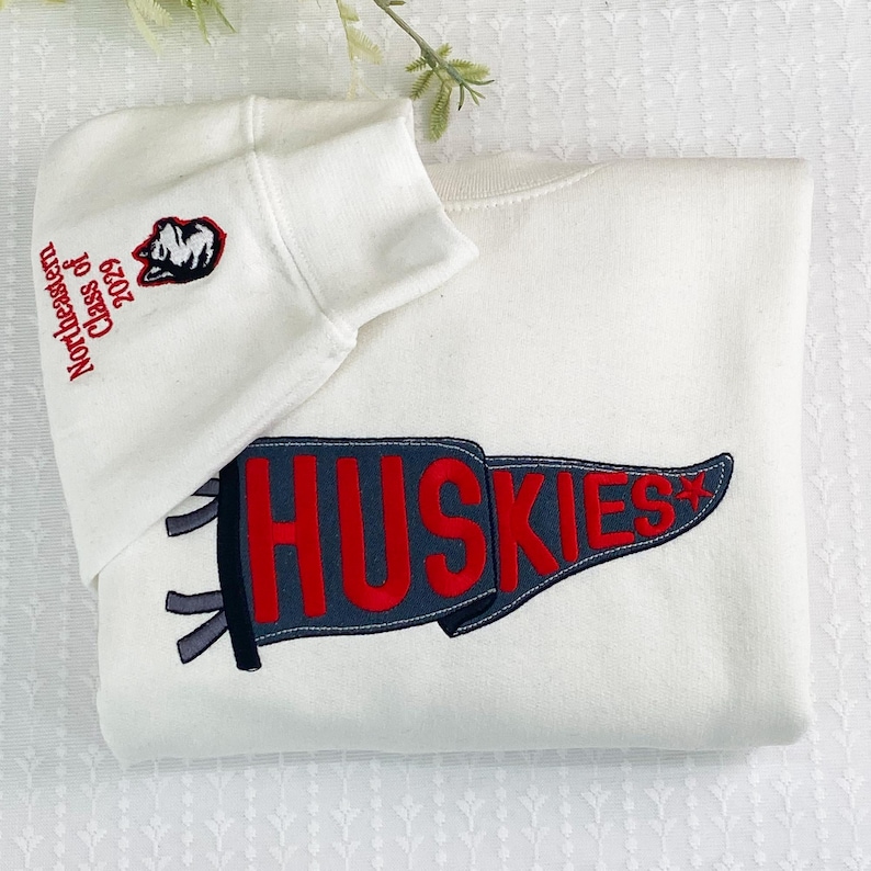 Puede incluir: Sudadera color crema con un dise&ntilde;o de bander&iacute;n "Huskies" rojo y azul marino. El pu&ntilde;o de la manga izquierda presenta el texto "Northeastern Class of 2029" y un logotipo de husky. La sudadera est&aacute; doblada cuidadosamente.