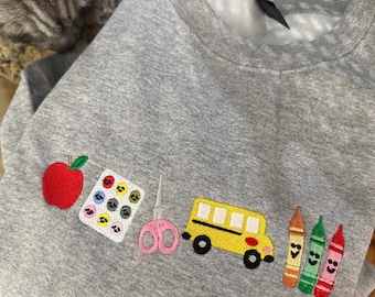 Op de afbeelding: Een grijs gemêleerd sweatshirt met ronde hals en geborduurde schoolthema-iconen op de borst, waaronder een rode appel, een verfpalet, een roze schaar, een gele schoolbus en drie kleurpotloden in oranje, groen en rood.