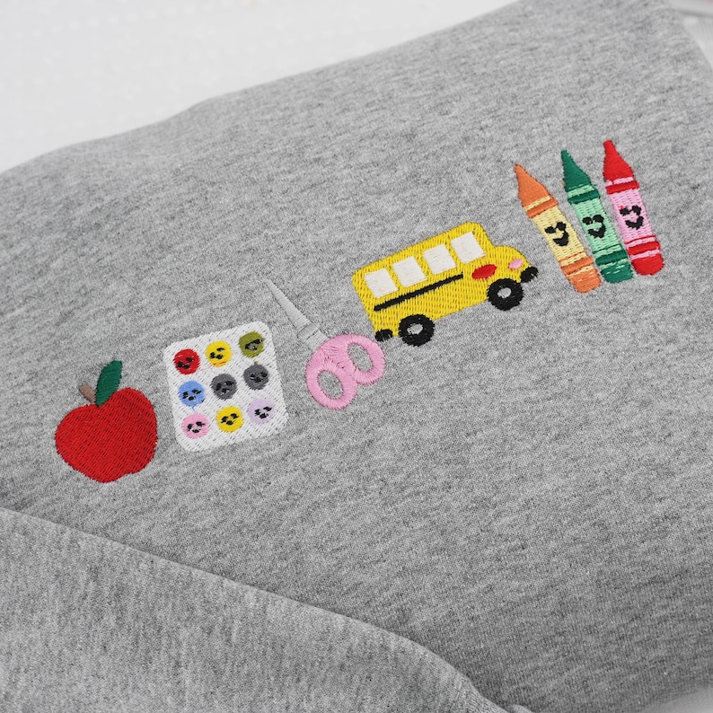 Folded grey sweatshirt with colorful embroidered school icons including apple, paint palette, scissors, school bus, and crayons – perfect back-to-school gift for teachers.