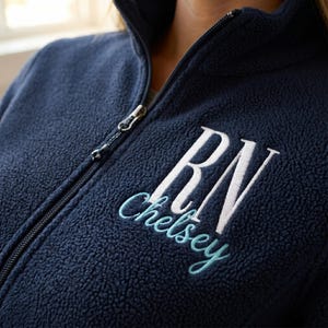 May include: Navy blue fleece jacket with a full zipper and a high collar. The jacket features the embroidered letters "RN" in white, with "Chelsey" embroidered in a light blue script font below.