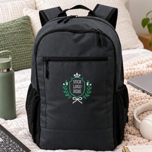 Könnte beinhalten: Ein anthrazitfarbener Rucksack mit Tragegriff, verstellbaren Schultergurten und zwei seitlichen Netztaschen. Auf der Vorderseite befindet sich ein grün-weißes gesticktes Wappenlogo mit dem Text YOUR LOGO HERE in einem dekorativen Lorbeerkranz.