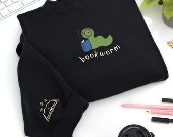 May include: Black long-sleeve shirt with a mock neck. The shirt features an embroidered green bookworm wearing glasses and holding a blue book, with the word "bookworm" below. The sleeve has an embroidered open book with stars.