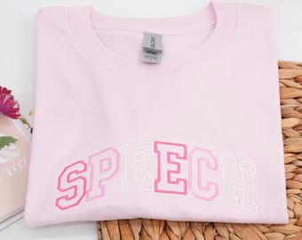 May include: A light pink sweatshirt with the word "SPEECH" embroidered across the front in shades of pink and white. The sweatshirt is folded neatly on a woven surface.