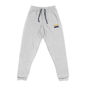 Puede incluir: Pantalones de chándal de color gris claro con cintura con cordón y un pequeño parche en la pierna izquierda. El parche presenta un diseño de rayas horizontales en morado, amarillo y negro. El cordón es azul marino.
