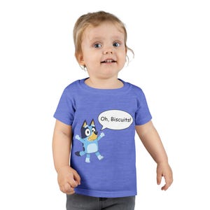 Op de afbeelding: Een blauw T-shirt met korte mouwen en een afbeelding van het tekenfilmfiguur Bluey met opgeheven armen en een tekstballon met de tekst Oh, Biscuits! Het shirt heeft een ronde hals en een gemêleerde textuur over de gehele stof.