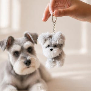 May include: A plush, gray and white Schnauzer dog keychain is held up in front of a real Schnauzer dog. The keychain is attached to a silver key ring. The dog is lying down on a white surface.