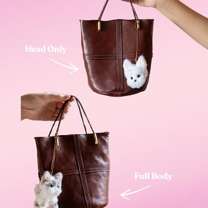 May include: Two brown leather tote bags with gold-toned hardware are displayed against a pink background. Each bag has a small, fluffy animal charm attached. One charm is a white dog, and the other is a gray and white cat.