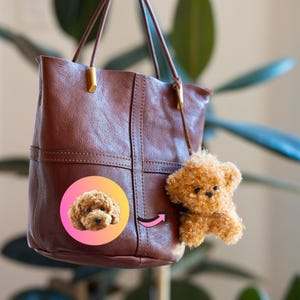 May include: A brown leather tote bag with a small, fluffy, light brown dog charm hanging from the handle. The bag has a stitched design and gold-colored hardware. A circular sticker with a dog's face is on the bag.