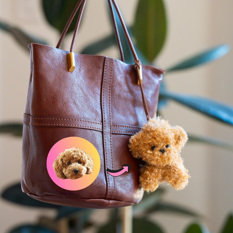 May include: A brown leather tote bag with a small, fluffy, light brown dog charm hanging from the handle. The bag has a stitched design and gold-colored hardware. A circular sticker with a dog's face is on the bag.