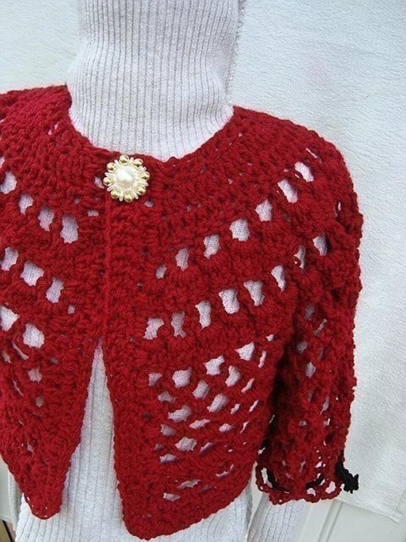 May include: Red crocheted bolero with a white pearl button. The bolero has a lacy design and is cropped.