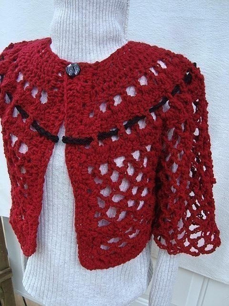 May include: Red crocheted shrug with black trim and a button closure. The shrug is made with a lacy stitch pattern.