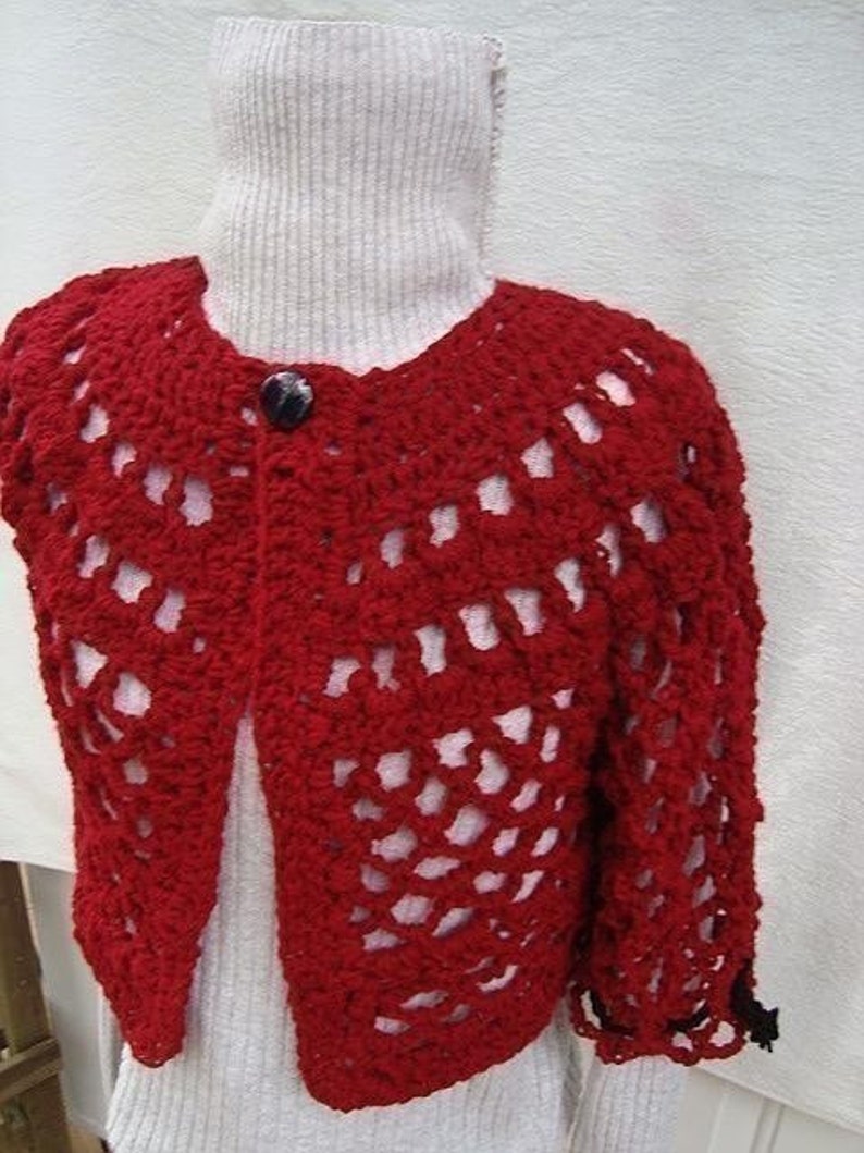 May include: Red crocheted cardigan with a button closure and a lacy pattern. The cardigan is shown on a white mannequin.