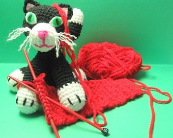 May include: A black and white crocheted cat with green eyes sits on a red crocheted blanket with red knitting needles and a ball of red yarn.