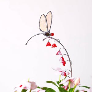 Op de afbeelding: Een decoratieve metalen tuinpaal met een libelle en een gebogen steel met rode bloemaccenten. De libelle heeft crèmekleurige vleugels en een bruin lichaam. De paal staat in een pot met roze en witte bloemen.