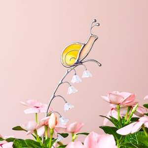 May include: A decorative garden stake featuring a stained glass snail with a yellow shell and cream-colored body. The snail sits atop a silver stem with white flower accents. Pink flowers and green leaves are in the foreground against a pink background.