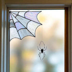 May include: A stained glass window hanging featuring a corner spider web design with iridescent white and purple glass panels held together by black metal solder lines, with a small black metal spider dangling from a thin wire below the web.