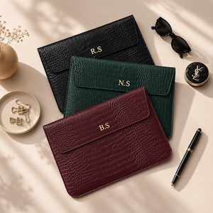 May include: Three personalized laptop sleeves in black, green, and burgundy, with gold initials. The sleeves have a textured, crocodile-like pattern. Accessories include sunglasses, a compact, and a pen.