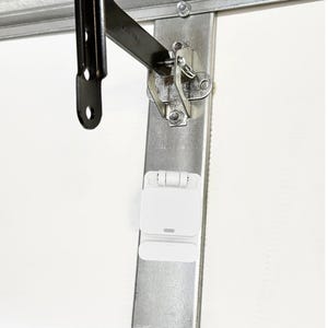 May include: A close-up of a garage door opener mechanism. The image shows a white, rectangular sensor attached to a metal track. A black metal arm and hinge are also visible, along with various nuts and bolts.