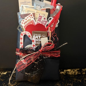 May include: A black gift package wrapped with twine and black lace, topped with a dark tag featuring stars. It contains a Trader Joe's oat chocolate bar, a red heart, stickers, a red tassel, a red pen, a white pen, and a library card stationery item.
