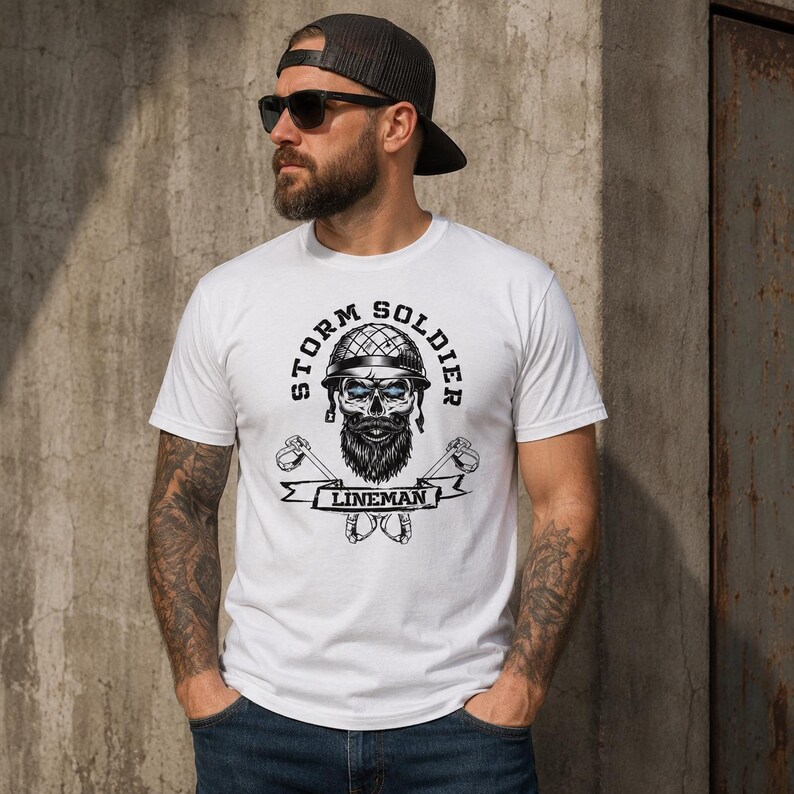 Op de afbeelding: Een wit t-shirt met korte mouwen voorzien van een afbeelding van een schedel met een helm en baard, met de tekst "STORM SOLDIER" in een boog erboven en "LINEMAN" op een banner eronder, geflankeerd door gekruiste gereedschappen in zwart, grijs en blauw.