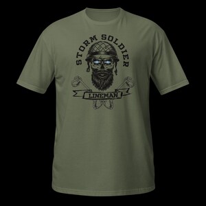Op de afbeelding: Een olijfgroen t-shirt met een grafische print van een bebaarde schedel die een helm in militaire stijl draagt. De tekst STORM SOLDIER staat in een boog boven de schedel en het woord LINEMAN staat op een banner eronder, geflankeerd door gekruiste gereedschappen.