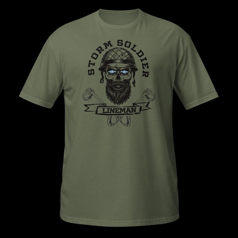 Op de afbeelding: Een olijfgroen t-shirt met een grafische print van een bebaarde schedel die een helm in militaire stijl draagt. De tekst STORM SOLDIER staat in een boog boven de schedel en het woord LINEMAN staat op een banner eronder, geflankeerd door gekruiste gereedschappen.