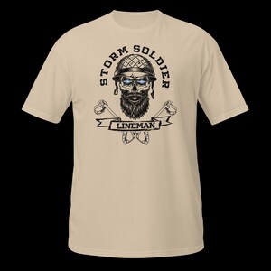 Op de afbeelding: Een beige t-shirt met korte mouwen en een grafische print van een schedel met een militaire helm en baard. De tekst STORM SOLDIER staat in een boog erboven, met LINEMAN op een banner eronder. Twee gekruiste klimhaken staan achter de banner.
