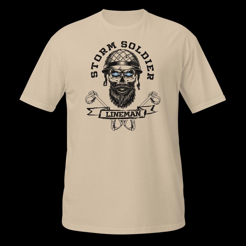 Op de afbeelding: Een beige t-shirt met korte mouwen en een grafische print van een schedel met een militaire helm en baard. De tekst STORM SOLDIER staat in een boog erboven, met LINEMAN op een banner eronder. Twee gekruiste klimhaken staan achter de banner.